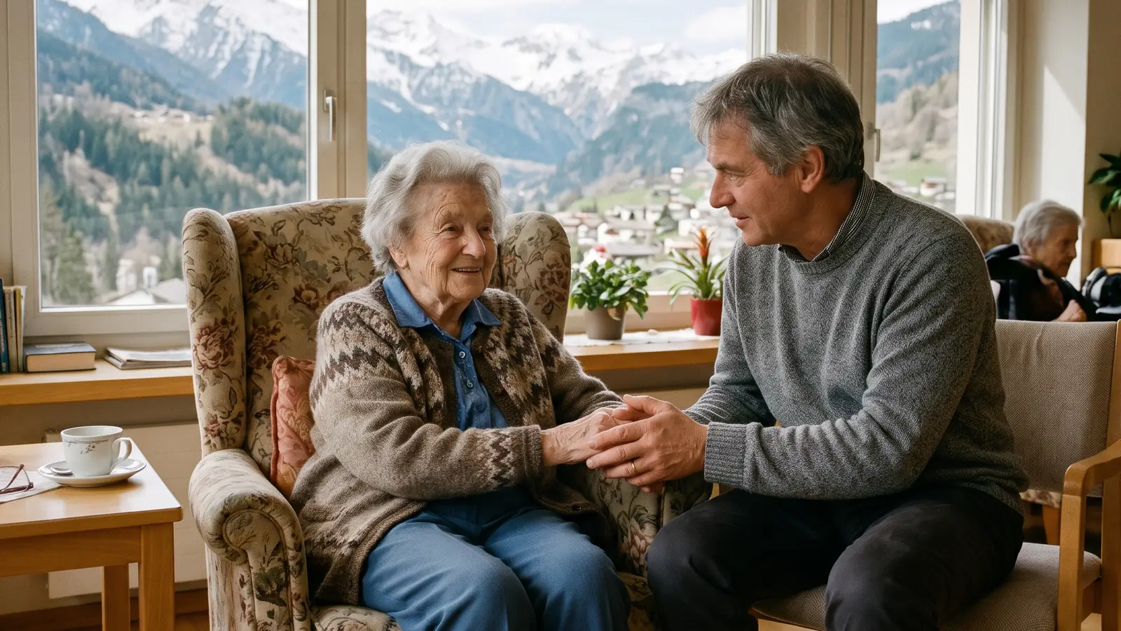 Une résidente très âgée dans un fauteuil d'EMS tient la main de son fils venu lui rendre visite, les montagnes derrière la fenêtre.